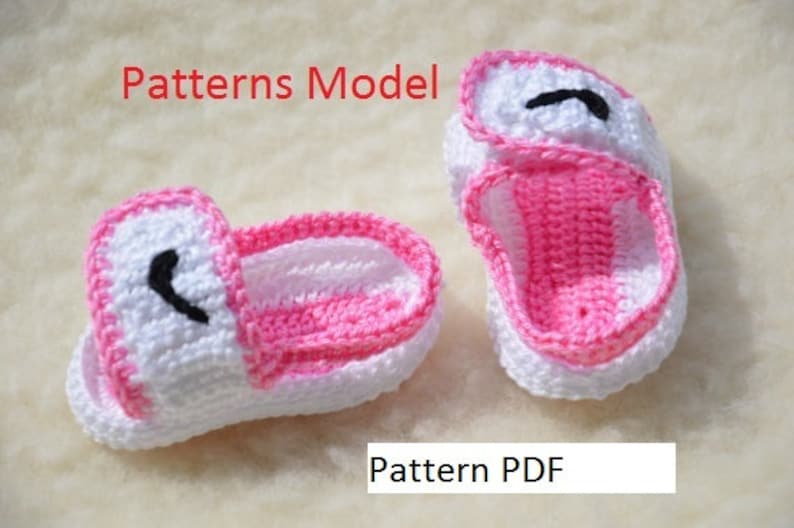 Puede incluir: Un par de zapatos de beb&eacute; de crochet blanco y rosa con un dise&ntilde;o de ojo negro. Los zapatos est&aacute;n colocados sobre una superficie blanca y peluda. El texto "Patterns Model" est&aacute; en rojo en la parte superior de la imagen. El texto "Pattern PDF" est&aacute; en negro en la parte inferior de la imagen.