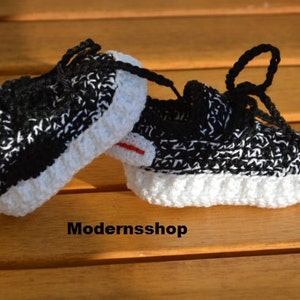 May include: A pair of black and white crocheted baby shoes with white laces. The shoes are in the shape of sneakers and have a white sole.