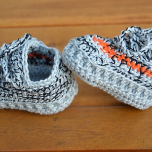 May include: A pair of gray crocheted baby booties with black and orange accents. The booties have a lace-up design.