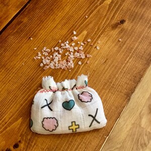 May include: A small white fabric pouch with a pink, blue, and yellow design. The pouch is filled with pink Himalayan salt crystals.
