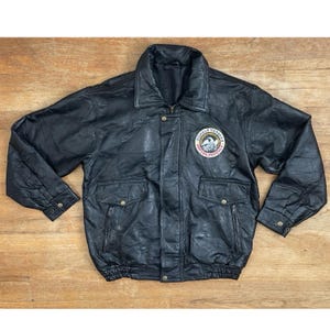 May include: Black leather jacket with a collared neckline, two front pockets, and a patch on the chest. The patch features text and an emblem. The jacket has a zipper closure and buttoned cuffs.