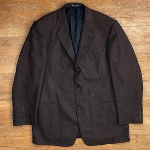 Può includere: Blazer marrone con un sottile motivo a quadri. La giacca ha un revers a lancia, tre bottoni e una tasca sul petto. La fodera interna è blu scuro. Il blazer è appoggiato su una superficie di legno.