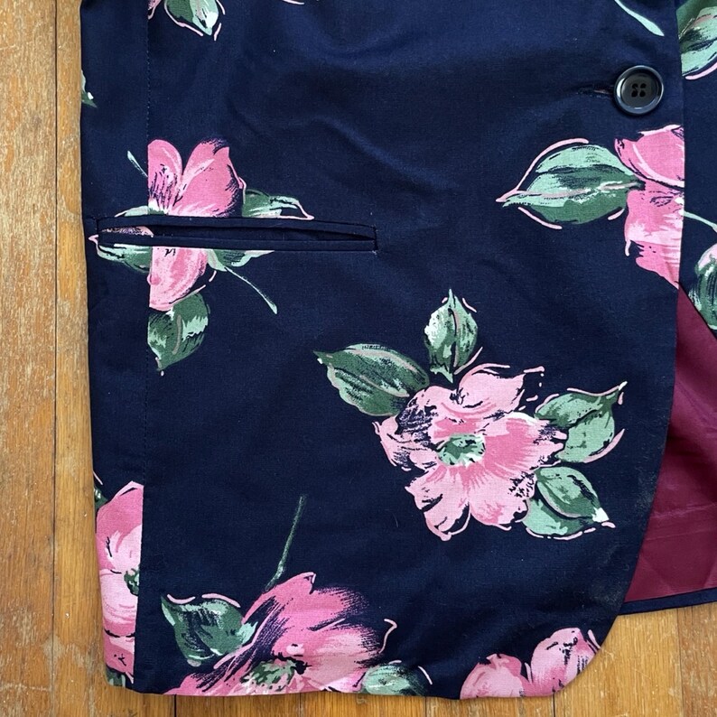 K&ouml;nnte beinhalten: Ein marineblauer Blazer mit einem Blumenmuster mit rosa Bl&uuml;ten und gr&uuml;nen Bl&auml;ttern. Der Blazer hat einen einzigen Knopfverschluss und eine Tasche auf der linken Seite.