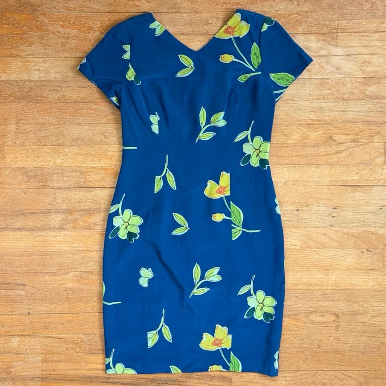 Puede incluir: Un vestido azul con cuello en V y mangas cortas. El vestido est&aacute; adornado con un estampado floral con flores y hojas amarillas y verdes. El vestido es aproximadamente hasta la rodilla.