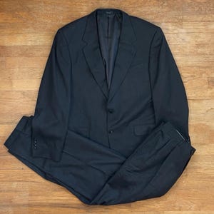 May include: A dark navy blue suit set, including a two-button blazer and matching trousers. The blazer has notched lapels and a chest pocket. The trousers have pleats and side pockets. The suit is laid out on a wooden surface.