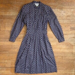 Può includere: Un vestito blu navy con un motivo ripetuto di piccoli cerchi colorati. L'abito ha maniche lunghe, un'abbottonatura frontale e una vita aderente. La gonna è a pieghe e l'abito arriva al ginocchio.