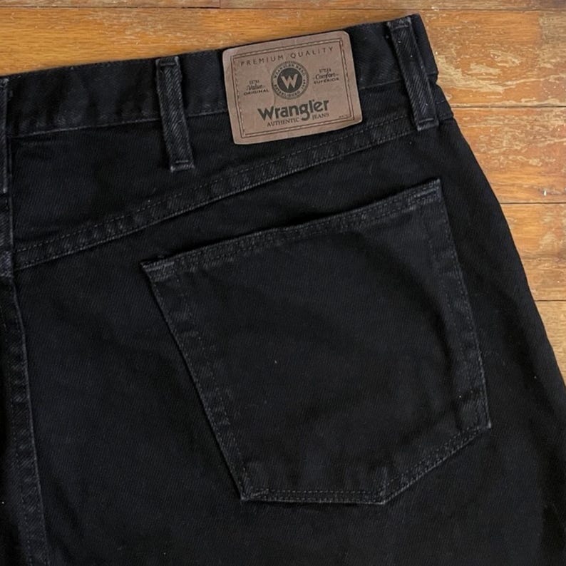 Puede incluir: Primer plano de unos vaqueros Wrangler negros. Los vaqueros tienen un dise&ntilde;o cl&aacute;sico de cinco bolsillos con un parche de cuero marr&oacute;n en la parte trasera que dice "Wrangler Authentic Jeans". Los vaqueros est&aacute;n hechos de un material de mezclilla duradero.