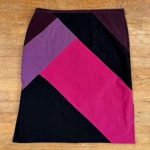 May include: A knee-length skirt with a colour block design. The skirt features panels of black, purple, magenta, and burgundy. The skirt is made of a smooth fabric and has a straight silhouette.