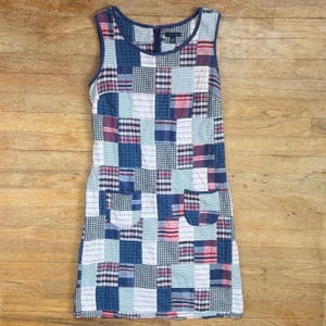 May include: Sleeveless patchwork dress featuring a mix of blue, red, and white squares. The dress has two front pockets and a zipper closure at the neckline. The dress is made of a quilted fabric and has a round neckline.