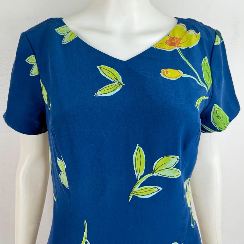 Puede incluir: Vestido azul marino con cuello en V y mangas cortas. El vestido est&aacute; adornado con un estampado floral con flores amarillas y hojas verdes. El dise&ntilde;o floral est&aacute; disperso por la tela.