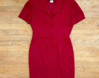 Könnte beinhalten: Ein einfarbig rotes Kleid mit kurzen Ärmeln, Reverskragen, einer durchgehenden Knopfleiste mit sieben passenden roten Knöpfen und zwei horizontalen Eingrifftaschen auf Taillenhöhe. Das Innenetikett trägt die Aufschrift Donna Ricco New York Petite.