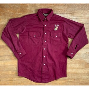 May include: A long-sleeved burgundy button-down shirt with a deer embroidered on the left chest. The shirt has two flap pockets on the front.