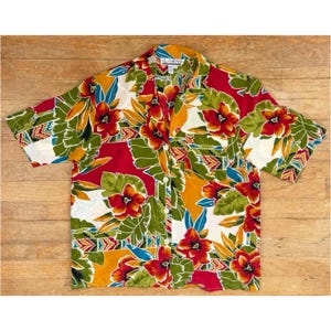 May include: A short-sleeved button-down shirt with a vibrant floral pattern. The shirt features red, orange, green, and white flowers and leaves against a red and yellow background. The shirt has a collar and is made of a lightweight fabric.