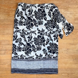 May include: A wrap skirt featuring a black floral pattern on a white background. The skirt includes a side tie closure and a hemline detailed with a horizontal band of fine black and white stripes and a textured, speckled gray pattern.