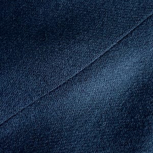 Op de afbeelding: Close-up van een getextureerde, marineblauwe stof met een diagonale naad. De stof heeft een licht versleten uiterlijk, met variaties in kleur en textuur. De afbeelding benadrukt de details van het materiaal.