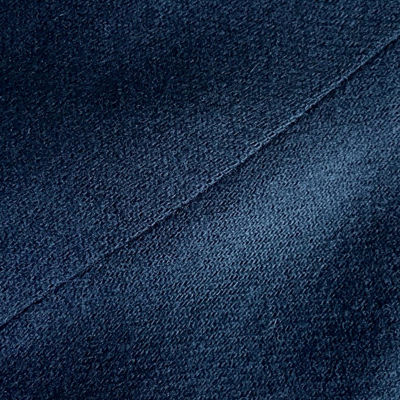 Op de afbeelding: Close-up van een getextureerde, marineblauwe stof met een diagonale naad. De stof heeft een licht versleten uiterlijk, met variaties in kleur en textuur. De afbeelding benadrukt de details van het materiaal.