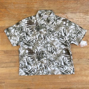 Puede incluir: Una camisa de manga corta con botones, fondo blanco y estampado tropical en tonos verdes y marrones. La camisa tiene cuello y cierre de botones.