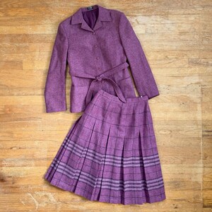 May include: A purple two-piece set consisting of a long-sleeve blazer and a pleated skirt. The blazer features a notched collar, a button-front closure, and a matching tie belt. The skirt has a dark purple and black plaid pattern throughout.