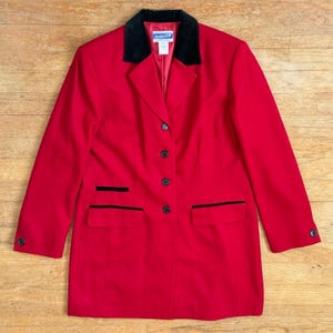 Può includere: Un blazer rosso acceso con colletto in velluto nero, quattro bottoni neri sul davanti e due tasche con patta bordate di nero. La giacca presenta maniche lunghe con un bottone nero su ogni polsino e un'etichetta del marchio Pendleton sul collo.