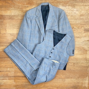 May include: A three-piece suit in a light blue plaid pattern. The suit includes a blazer, vest, and trousers. The blazer has a two-button closure and the vest has a button-down front. The suit is made of a woven fabric.