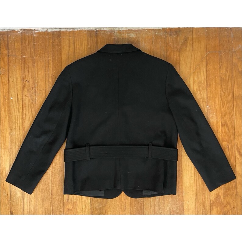 Op de afbeelding: Een zwarte blazer met een revers en een ceintuur in de taille. De jas heeft lange mouwen en een klassiek, getailleerd ontwerp. De blazer is gemaakt van een effen zwarte stof.