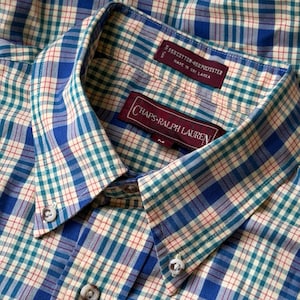 Peut inclure: Une chemise à col boutonné avec un motif à carreaux composé de lignes bleues, bleu sarcelle et rouges sur fond crème. Le col présente deux boutons visibles. Une étiquette indique Chaps Ralph Lauren taille M, 55% coton, 45% polyester.