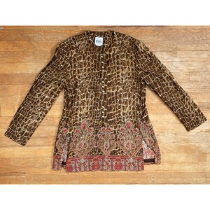 Peut inclure: Une chemise boutonnée à manches longues avec un motif marron et beige semblable à celui d'un reptile. L'ourlet inférieur présente un motif floral décoratif rouge, rose et or. La chemise est posée à plat sur une surface en bois.