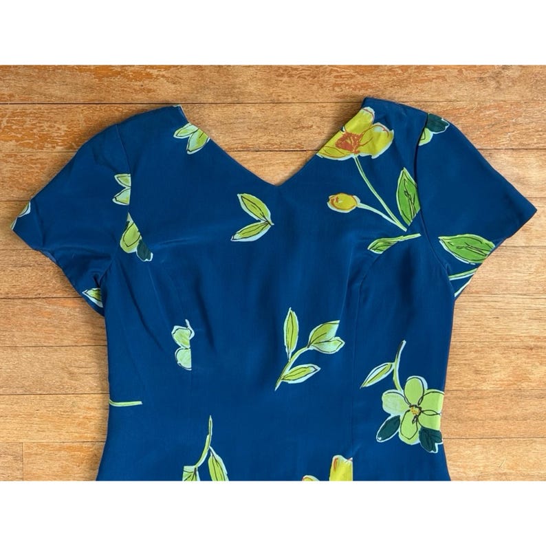 Puede incluir: Vestido azul marino con cuello en V y mangas cortas. El vestido est&aacute; adornado con un estampado floral con flores y hojas amarillas y verdes. El vestido est&aacute; hecho de una tela suave, posiblemente similar a la seda.