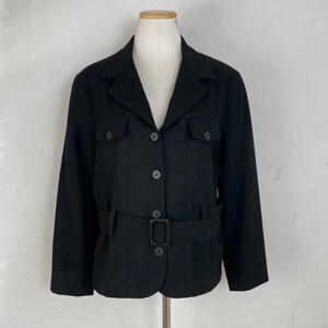 Op de afbeelding: Zwarte jas met een reverskraag, twee borstzakken en een bijpassende riem. De jas heeft een knoopsluiting aan de voorkant en wordt op een mannequin gepresenteerd. De jas is ongeveer 63,5 cm lang.