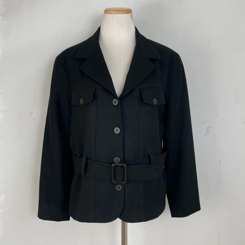 Op de afbeelding: Zwarte jas met een reverskraag, twee borstzakken en een bijpassende riem. De jas heeft een knoopsluiting aan de voorkant en wordt op een mannequin gepresenteerd. De jas is ongeveer 63,5 cm lang.