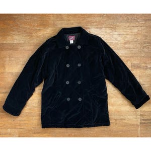 May include: A black velvet double-breasted jacket with a quilted pattern. The jacket has a notched collar, two rows of buttons, and two front pockets. The jacket is displayed on a wooden surface.