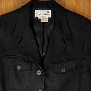 Op de afbeelding: Zwarte blazer met een klassiek ontwerp, met een reverskraag, twee voorzakken en decoratieve knopen. Het label luidt "Stanley Blacker For Gantos". De blazer is maat 6.