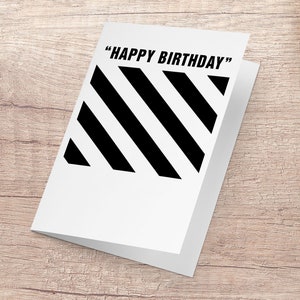 May include: White folded birthday card with black diagonal stripes and the text "HAPPY BIRTHDAY" in black. The card is on a light brown wooden surface.