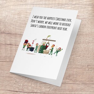 Puede incluir: Una tarjeta de Navidad con una ilustración de dibujos animados de duendes trabajando en una fábrica. La tarjeta dice "Te deseo la Navidad más feliz de todas, no te preocupes, trabajaremos para reducir la huella de carbono de Santa Claus el próximo año."