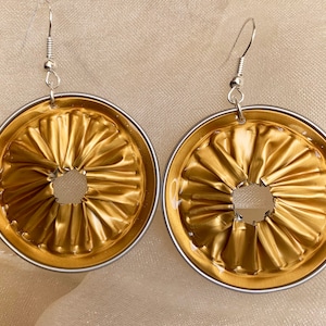 Puede incluir: Un par de pendientes de color dorado hechos con cápsulas de café reutilizadas. Los pendientes tienen una forma circular con un pequeño agujero en el centro. Los pendientes están unidos a ganchos plateados.