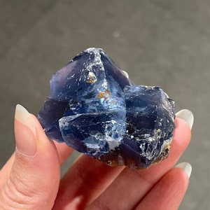 Pode incluir: Um aglomerado de cristais de fluorita azul com um toque de roxo e inclusões douradas. Os cristais são facetados e têm uma aparência translúcida.