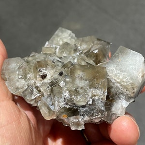 Pode incluir: Um aglomerado de cristais de fluorita cinzentos com um brilho ligeiramente iridescente. Os cristais est&atilde;o intercrescidos e t&ecirc;m faces afiadas e bem definidas.