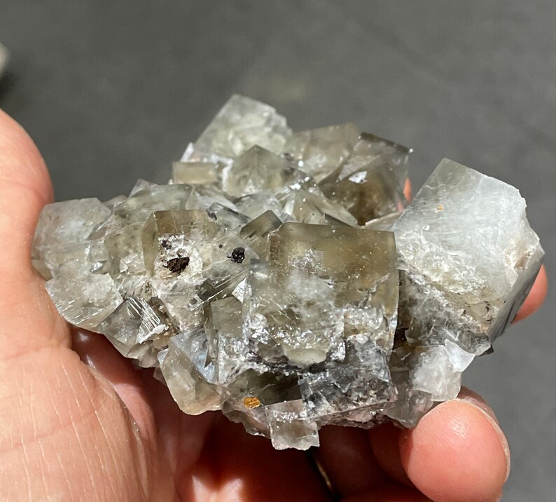 Pode incluir: Um aglomerado de cristais de fluorita cinzentos com um brilho ligeiramente iridescente. Os cristais est&atilde;o intercrescidos e t&ecirc;m faces afiadas e bem definidas.