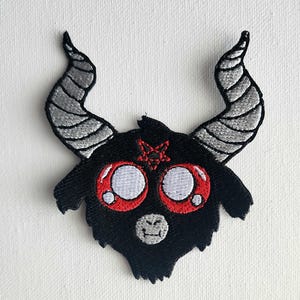 Pode incluir: Patch bordado com uma cabeça de cabra preta de desenho animado com olhos grandes com bordas vermelhas e uma estrela vermelha na testa. A cabra tem chifres cinzentos e uma boca pequena e sorridente. Para roupas ou acessórios.