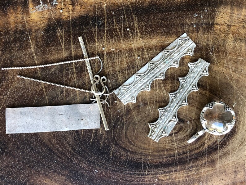 May include: A collection of silver jewelry making supplies on a wooden surface. The supplies include a silver chain, a silver bar, a silver piece with a decorative pattern, and a silver piece with a clear glass insert.