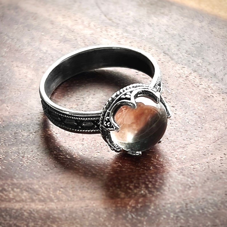 May include: A silver ring with a clear gemstone set in a detailed silver bezel. The ring band is decorated with a repeating pattern.