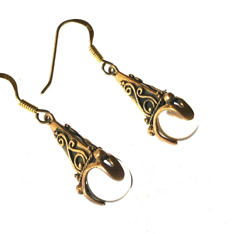 Puede incluir: Un par de pendientes de tono dorado con piedras de cristal transparente en forma de l&aacute;grima. Los pendientes tienen un dise&ntilde;o de filigrana.