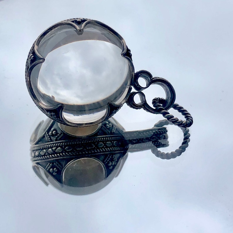 May include: A silver pendant featuring a large, clear crystal at its centre. The crystal is surrounded by intricate silver designs. The pendant is attached to a silver chain.