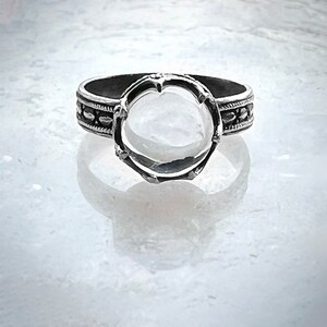 May include: A silver ring with a bezel setting for a round stone. The ring band is decorated with a beaded design.