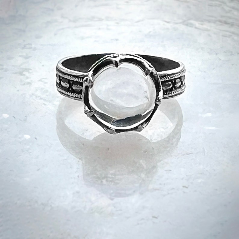 May include: A silver ring with a bezel setting for a round stone. The ring band is decorated with a beaded design.
