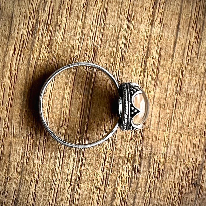 May include: A silver ring with a clear stone set in a silver bezel. The bezel is decorated with a detailed design.