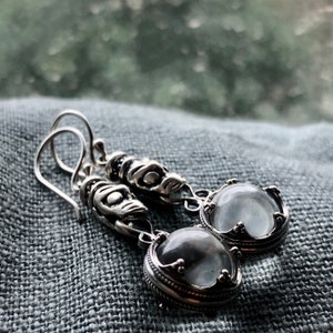 May include: Silver dangle earrings with clear round stones set in ornate silver frames. The earrings feature intricate silver designs.