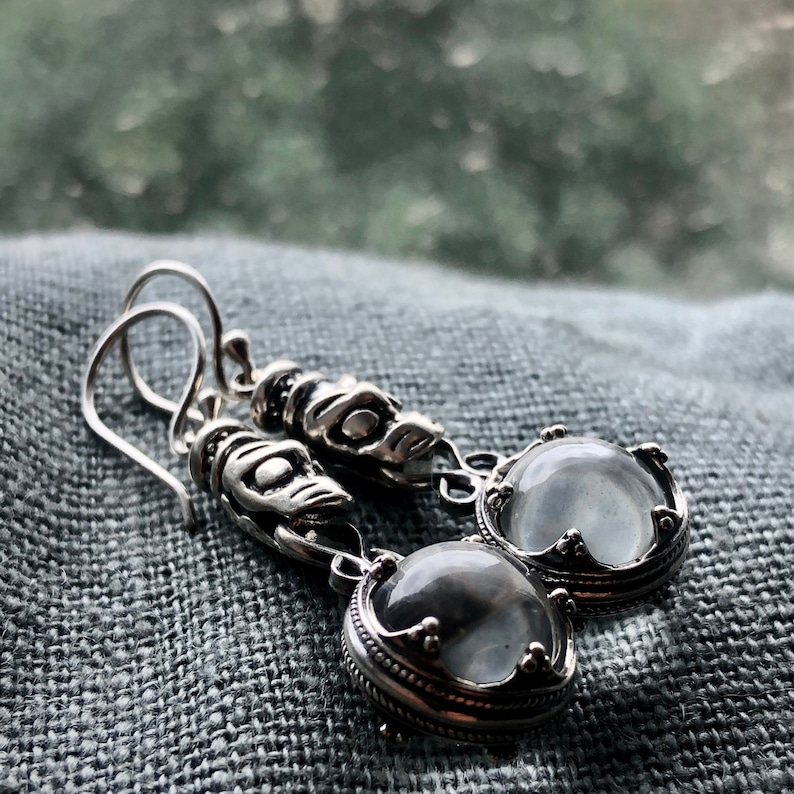 May include: Silver dangle earrings with clear round stones set in ornate silver frames. The earrings feature intricate silver designs.