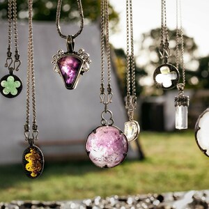 May include: A collection of silver necklaces with various gemstone pendants, including green, purple, and clear crystals. The necklaces are hanging from a string and the background is blurred.