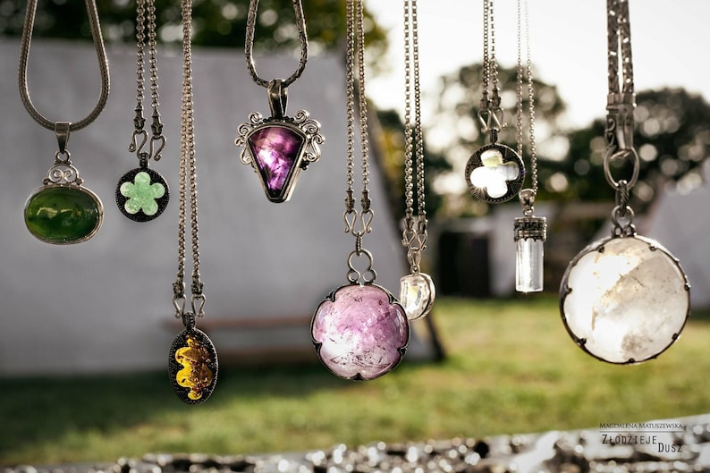May include: A collection of silver necklaces with various gemstone pendants, including green, purple, and clear crystals. The necklaces are hanging from a string and the background is blurred.